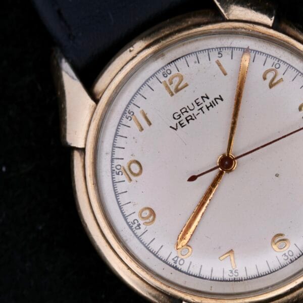 Close-up of a vintage Gruen Veri-Thin Fancy Lugs watch with a gold case and white dial. This elegant timepiece features gold hour markers and hands, capturing the time at about 10:09 against a dark background.
