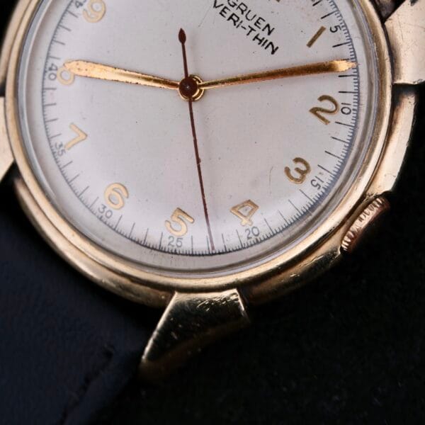 Close-up of a vintage Gruen Veri-Thin Fancy Lugs watch featuring a gold-toned case and white face. It has gold hands, classic numerals, and a partially visible black strap. The brand name is printed near the top of the dial.