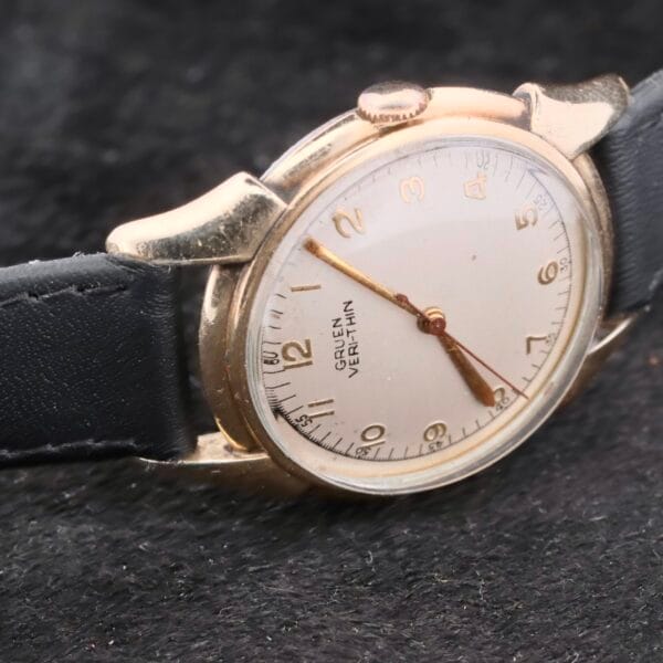 A vintage Gruen Veri-Thin Fancy Lugs wristwatch with a round gold-toned case and black leather strap features gold hour markers and hands on a white dial against a textured black background.