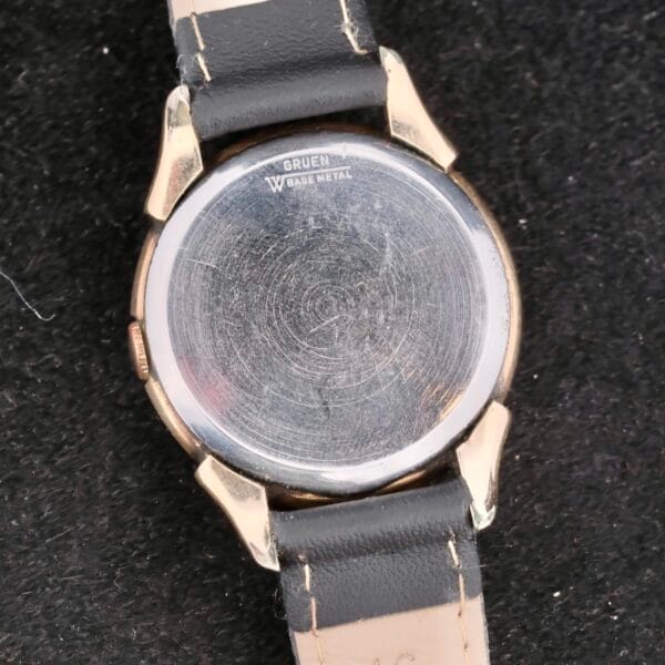 The image highlights the base metal case back of a Gruen Veri-Thin Fancy Lugs wristwatch, adorned with visible scratches. Its black leather strap elegantly rests on a textured black surface, embodying the timeless essence of this classic timepiece.