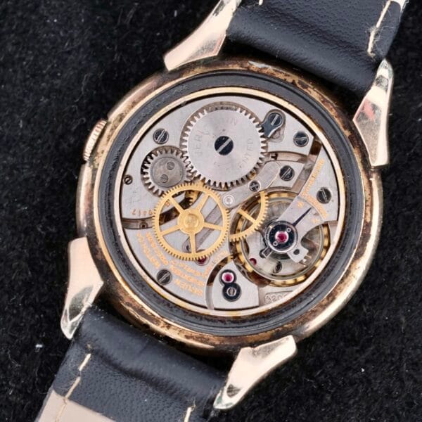 Close-up view of a vintage Gruen Veri-Thin Fancy Lugs wristwatch with a black leather strap, showcasing its intricate mechanical gears and components against a textured black surface.