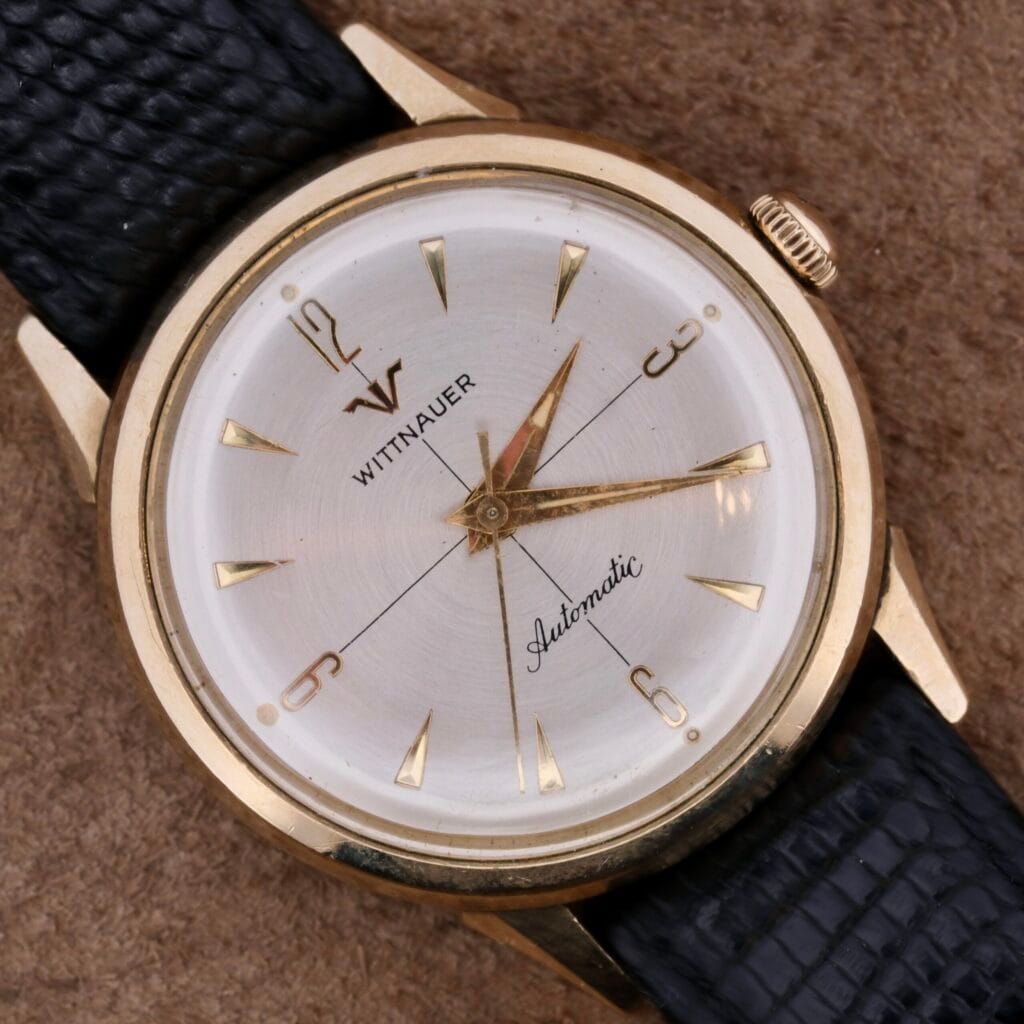 The Longines-Wittnauer Automatic wristwatch boasts a gold-tone case, white dial, and gold hour markers with a black textured leather strap, offering simple elegance.