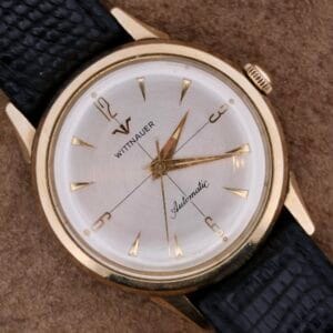 The Longines-Wittnauer Automatic wristwatch boasts a gold-tone case, white dial, and gold hour markers with a black textured leather strap, offering simple elegance.