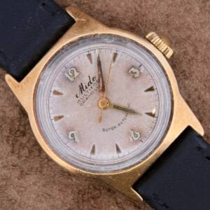 A vintage Mido Multifort Grand Luxe Super Automatic wristwatch with a gold-tone case and leather strap displays Arabic numerals and the labels Mido, MULTIFORT, and SUPER-AUTOMATIC, elegantly resting on a textured brown surface.