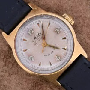 A vintage Mido Multifort Grand Luxe Super Automatic wristwatch with a gold-tone case and leather strap displays Arabic numerals and the labels Mido, MULTIFORT, and SUPER-AUTOMATIC, elegantly resting on a textured brown surface.