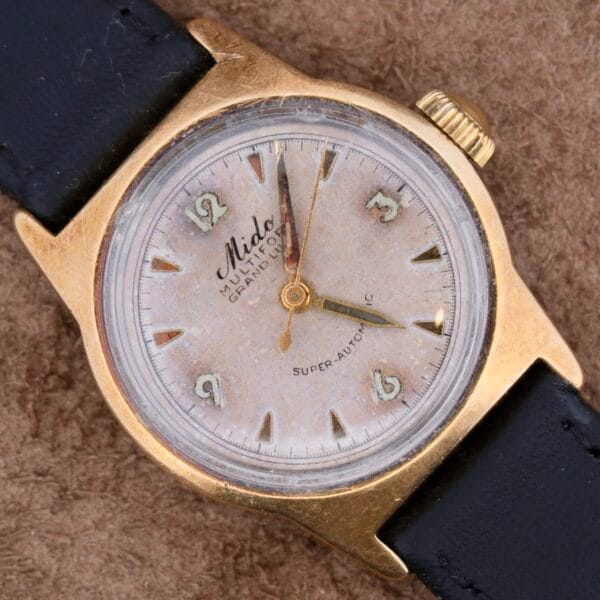 A vintage Mido Multifort Grand Luxe Super Automatic wristwatch with a gold-tone case and leather strap displays Arabic numerals and the labels Mido, MULTIFORT, and SUPER-AUTOMATIC, elegantly resting on a textured brown surface.
