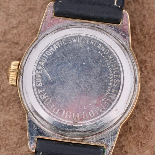 The image shows the scratched back of a vintage wristwatch with a black strap. The engraving reads AUTOMATIC SWITZERLAND STAINLESS STEEL and Mido Multifort Grand Luxe Super Automatic. The watch lies on a textured brown surface.