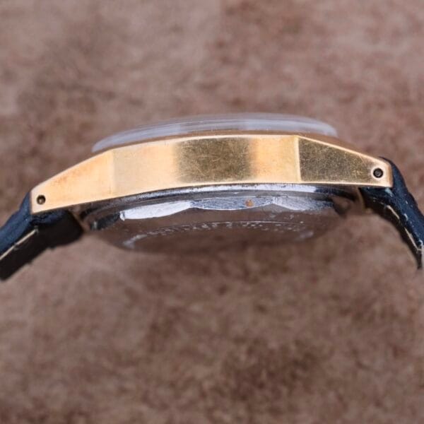 A side view of the Mido Multifort Grand Luxe Super Automatic wristwatch showcases its gold-tone case, crystal dome, and black leather strap as it rests on a textured beige surface.
