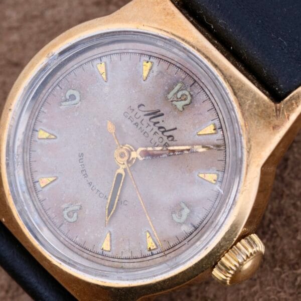 Close-up of a vintage Mido Multifort Grand Luxe Super Automatic watch with a gold case and faded dial. It features numerals, triangular markers, two hands, and a sweep second hand, exuding aged charm. A partially visible black leather strap adds elegance.