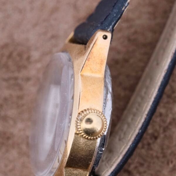 Close-up of a vintage Mido Multifort Grand Luxe Super Automatic wristwatch with a gold metal case and black leather strap. The crown is visible with detailed gear-like features against a blurred beige background.
