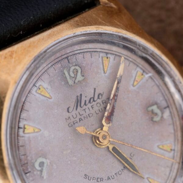 A close-up of a vintage gold wristwatch reveals Mido Multifort Grand Luxe Super Automatic on its worn, round face. The tarnished hour markers and hands echo past grandeur, while the distinctive wear on its outer edge adds to its antique allure.