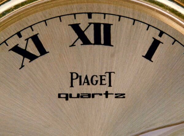 Close-up of the Piaget Quartz 18k Gold Classic watch face with Roman numerals XI, XII, and I. The words Piaget and quartz are elegantly printed in black on its brushed metal surface, reflecting a classic and sophisticated style.