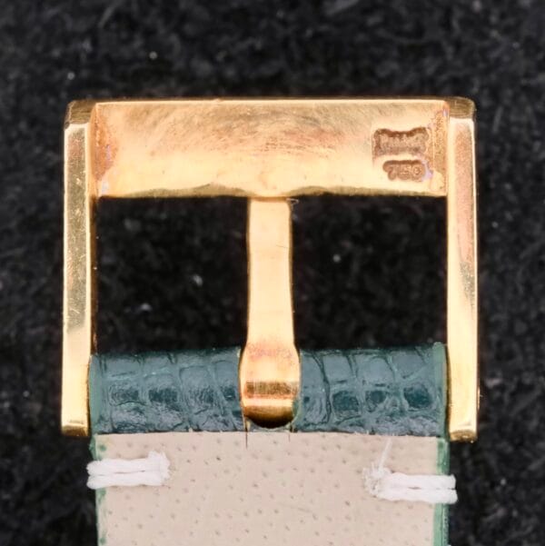 Close-up of a Piaget Quartz 18k Gold Classic watch buckle on a green textured leather strap with white stitching, elegantly engraved and resting against a black background.