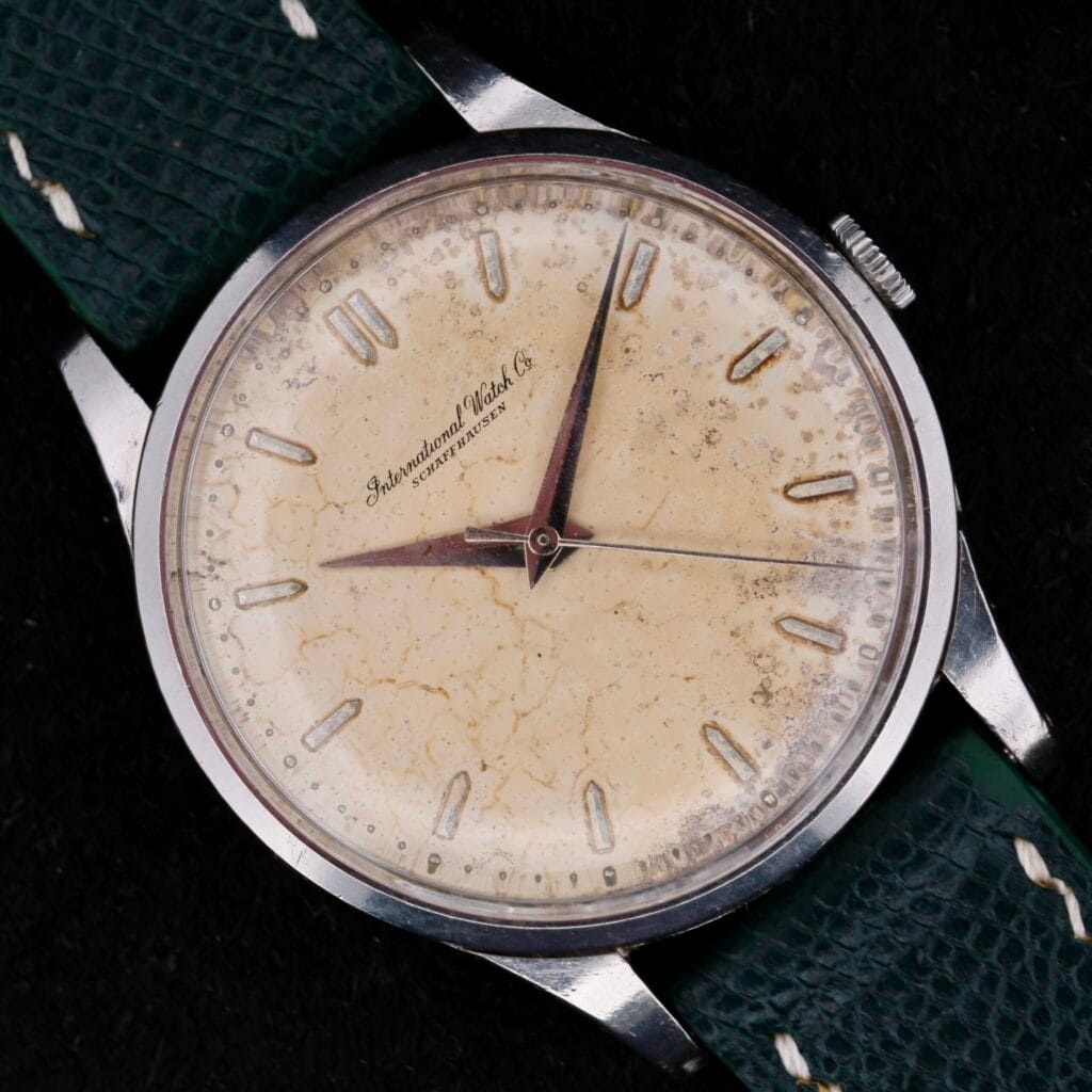 The IWC Caltrava Jumbo Cal. 89 Stainless Steel watch boasts an aged cream dial with markers and hands, paired with a stainless steel case and a dark green leather strap. The patina adds to its timeless appeal.