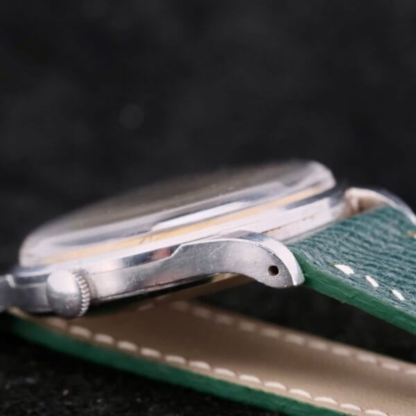 Close-up of an IWC Caltrava Jumbo Cal. 89 Stainless Steel wristwatch with a metallic case and a green leather strap, laid flat to showcase the side and textured strap against a dark background.