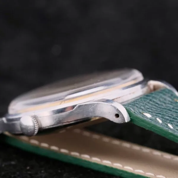 Close-up of an IWC Caltrava Jumbo Cal. 89 Stainless Steel wristwatch with a metallic case and a green leather strap, laid flat to showcase the side and textured strap against a dark background.