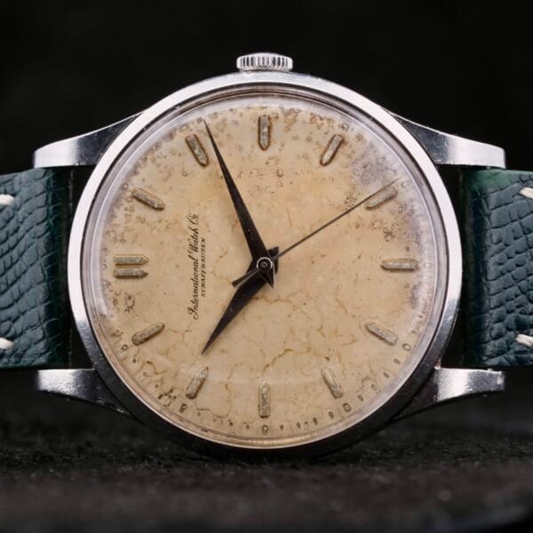 The IWC Caltrava Jumbo Cal. 89 Stainless Steel wristwatch boasts a worn, cream-colored dial with black markers and hands, all encased in metal. Its distressed green leather strap enhances its vintage appeal, while the partially visible brand name on the dial adds timeless elegance.