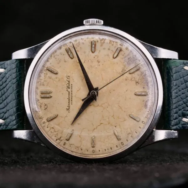 The IWC Caltrava Jumbo Cal. 89 Stainless Steel wristwatch boasts a worn, cream-colored dial with black markers and hands, all encased in metal. Its distressed green leather strap enhances its vintage appeal, while the partially visible brand name on the dial adds timeless elegance.