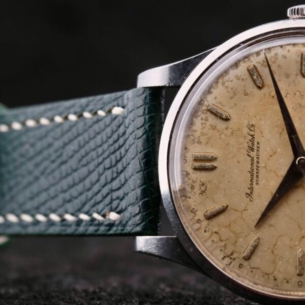 Close-up of an IWC Caltrava Jumbo Cal. 89 Stainless Steel wristwatch, showcasing a weathered beige dial with thin black hour markers and intricate hands, accented by a textured aged background and a dark, worn leather strap.