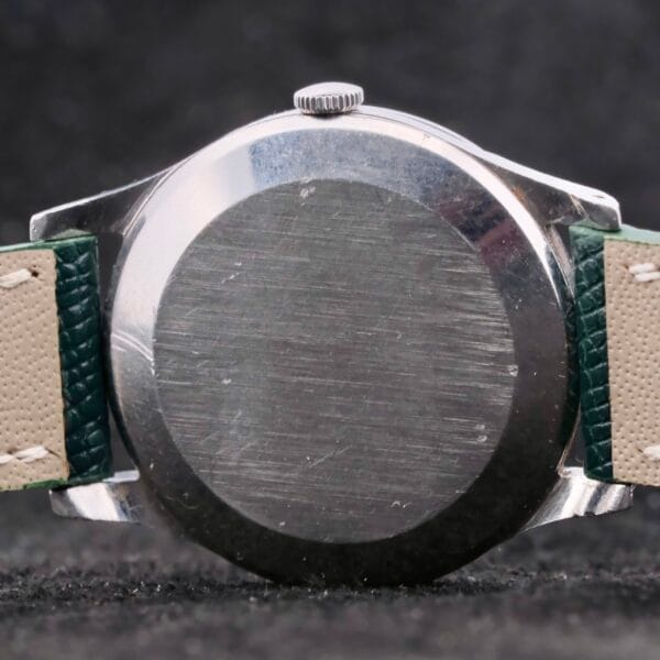 A close-up of the IWC Caltrava Jumbo Cal. 89 Stainless Steel wristwatch showcases its metallic allure, highlighted by a green and beige leather strap. The surface displays a scratched, worn texture against a dark backdrop.