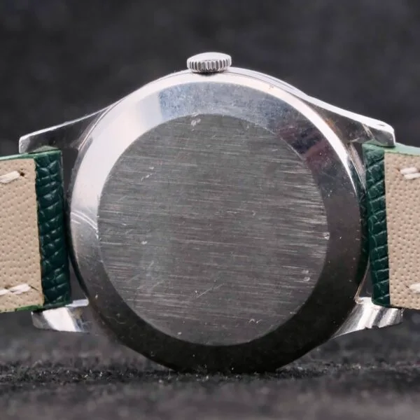 A close-up of the IWC Caltrava Jumbo Cal. 89 Stainless Steel wristwatch showcases its metallic allure, highlighted by a green and beige leather strap. The surface displays a scratched, worn texture against a dark backdrop.