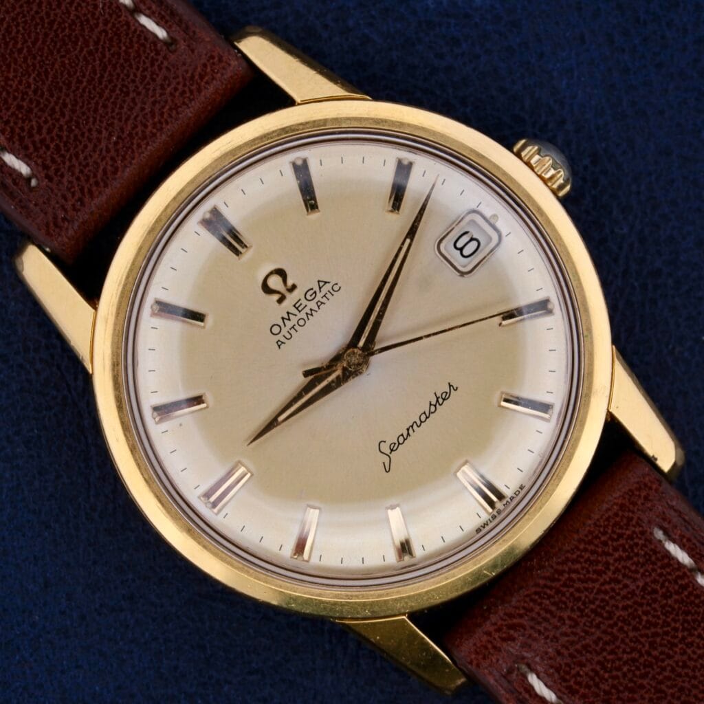 A close-up of an Omega Seamaster Date Automatic Cal 565 watch features a gold-colored case, cream dial with silver hour markers, a date window at 3 oclock, and Seamaster in cursive on a dark blue background, complemented by a brown leather strap.