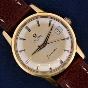 A close-up of an Omega Seamaster Date Automatic Cal 565 watch features a gold-colored case, cream dial with silver hour markers, a date window at 3 oclock, and Seamaster in cursive on a dark blue background, complemented by a brown leather strap.