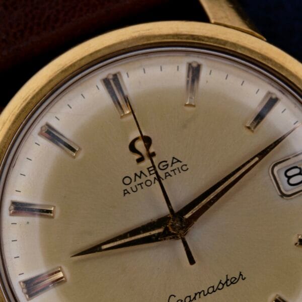 Close-up of a vintage Omega Seamaster Date Automatic Cal 565 watch face with gold hour markers, black hands, and the iconic Omega logo above Automatic. The date window displays the number 8.