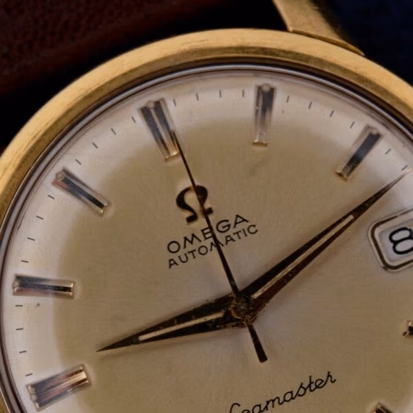 Close-up of a vintage Omega Seamaster Date Automatic Cal 565 watch face with gold hour markers, black hands, and the iconic Omega logo above Automatic. The date window displays the number 8.