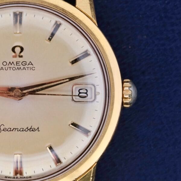 Close-up of a vintage Omega Seamaster Date Automatic Cal 565 watch with a gold case, white dial, and date window set to the 8th. This elegant timepiece boasts silver hour markers and hands against a captivating blue background.