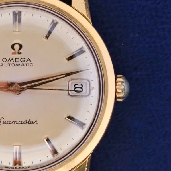 Close-up of a vintage Omega Seamaster Date Automatic Cal 565 watch with a gold case, white dial, and date window set to the 8th. This elegant timepiece boasts silver hour markers and hands against a captivating blue background.