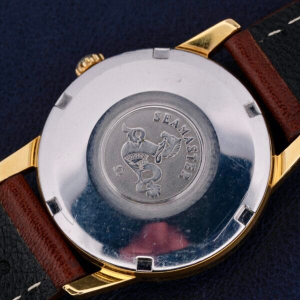 Close-up of the Omega Seamaster Date Automatic Cal 565s back, with Seamaster and Date finely engraved on a silver surface and an emblem. The watch features a gold edge, a two-tone brown and black leather strap, set against a dark backdrop.