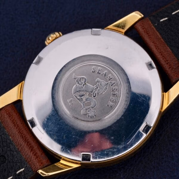 Close-up of the Omega Seamaster Date Automatic Cal 565s back, with Seamaster and Date finely engraved on a silver surface and an emblem. The watch features a gold edge, a two-tone brown and black leather strap, set against a dark backdrop.