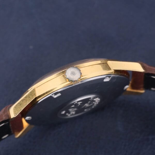 A side view of the Omega Seamaster Date Automatic Cal 565 showcases a gold-toned case, round crown, dark face, and brown leather strap, elegantly set against a dark blue background.