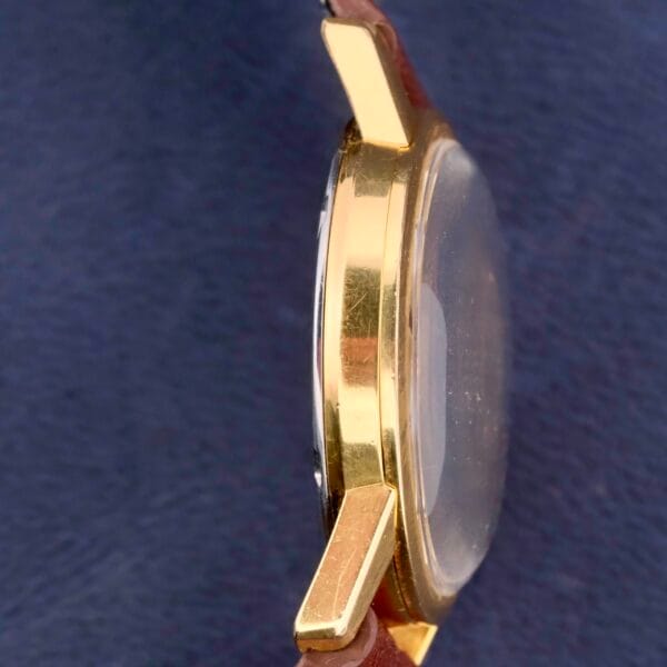 The Omega Seamaster Date Automatic Cal 565 showcases a gold case with a brown leather strap, elegantly set against a dark blue fabric background.