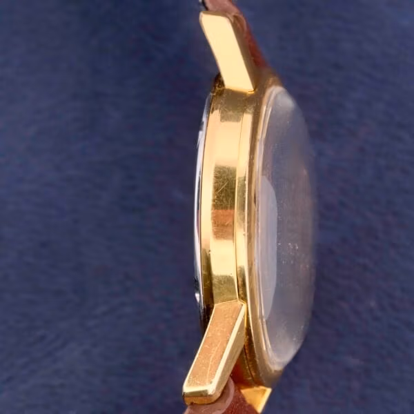 The Omega Seamaster Date Automatic Cal 565 showcases a gold case with a brown leather strap, elegantly set against a dark blue fabric background.