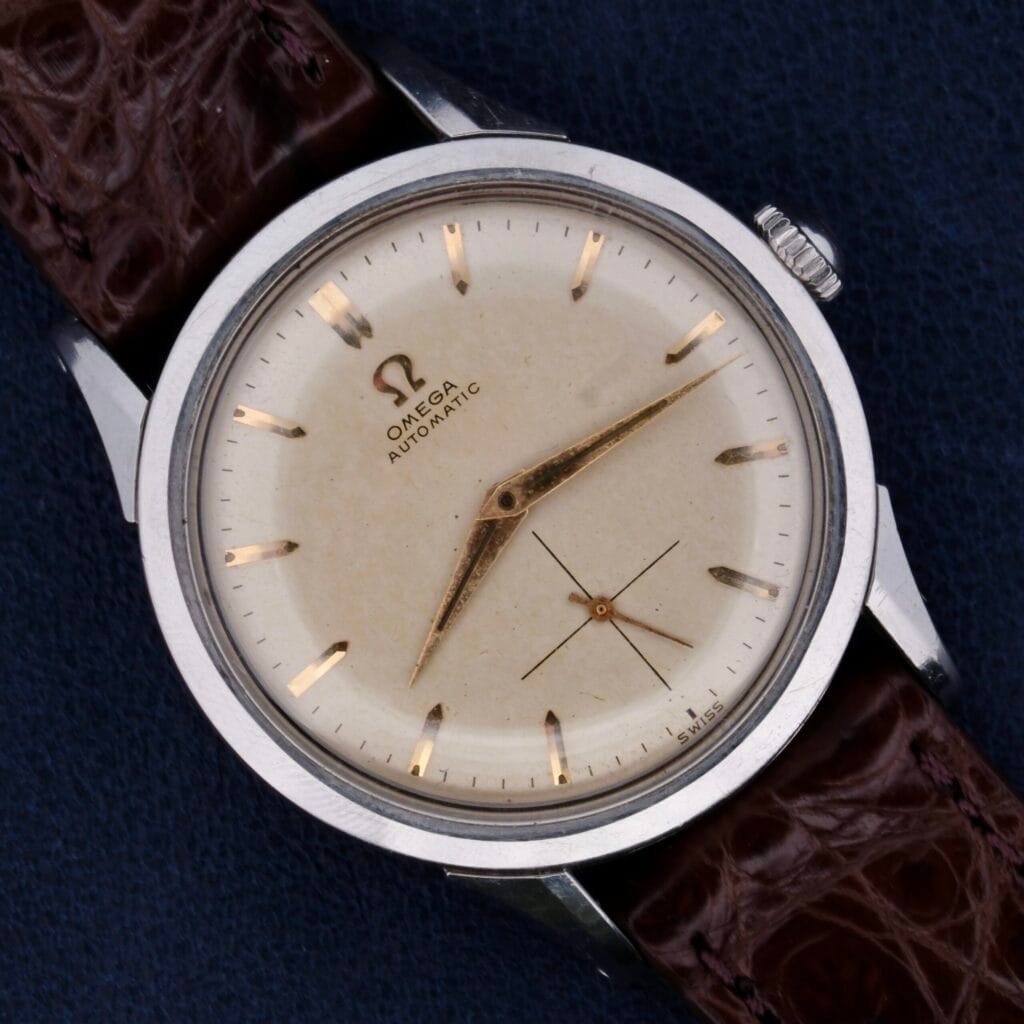 An Omega Automatic Bumper Cal 344 Stainless Steel watch with a cream dial and silver hour markers is displayed on a dark blue surface. It features thin, elegant hands and a brown leather strap, exuding classic and timeless appeal.