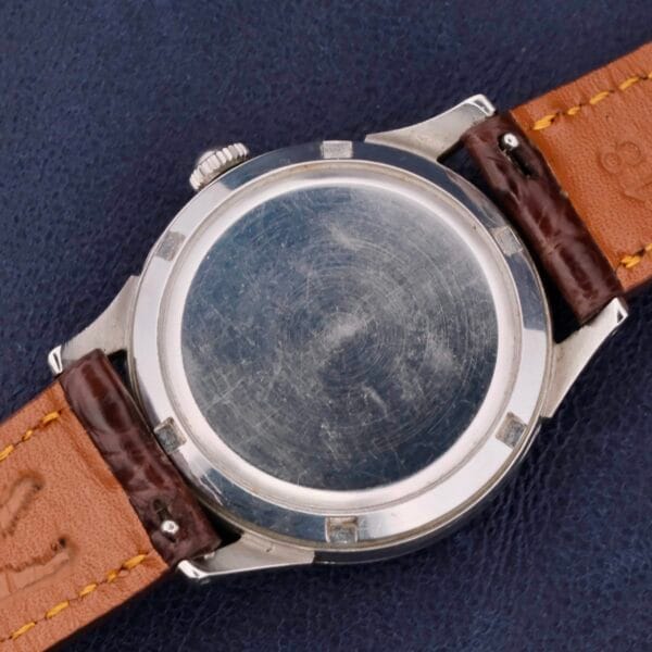 The image shows the back of an Omega Automatic Bumper Cal 344 Stainless Steel watch, with a silver metal case and brown leather straps, resting on a dark blue surface. The watch back is plain and slightly scratched.