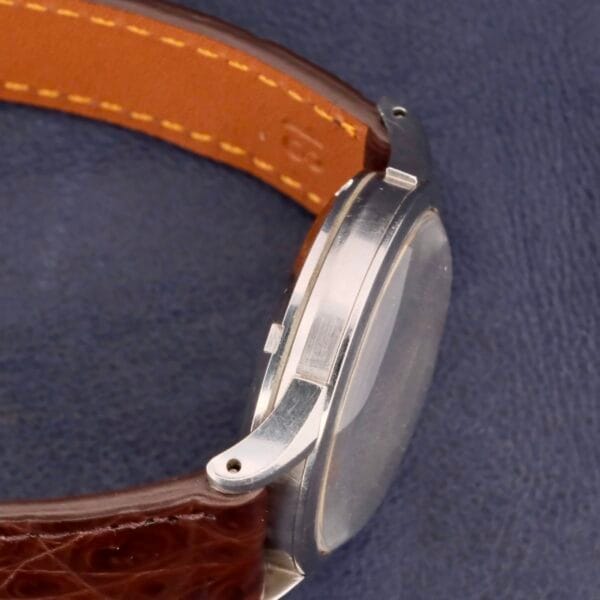 Close-up of an Omega Automatic Bumper Cal 344 Stainless Steel watch with a brown leather strap, on a dark blue surface. The side view showcases its slim profile and detailed stitching.