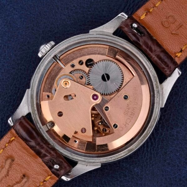 A close-up of a luxury Omega Automatic Bumper Cal 344 Stainless Steel Watch reveals its intricate mechanical movement through an open case back. The watch features a brown leather strap and metallic-finished interior gears, set against a dark, textured background.