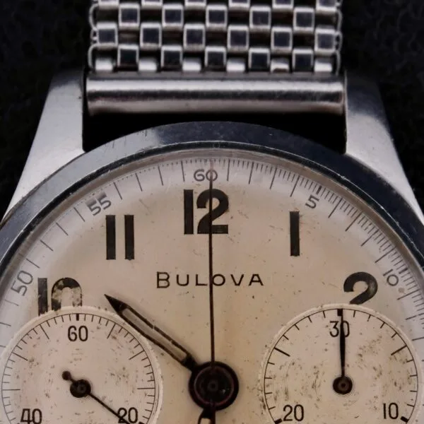 Close-up of a vintage Bulova Chronograph 13AK Valjoux 23 Stainless Steel watch with a silver mesh band, featuring two subdials and black numerals. The time is set to 12:00 against a textured black background.