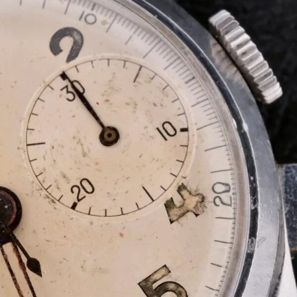 A close-up of the Bulova Chronograph 13AK Valjoux 23 watch face reveals its vintage charm. The weathered dial features a small seconds sub-dial at 9 oclock and visible scratches, while elegant hands point between 2 and 3. Its timeless look is completed by bold black numbers.