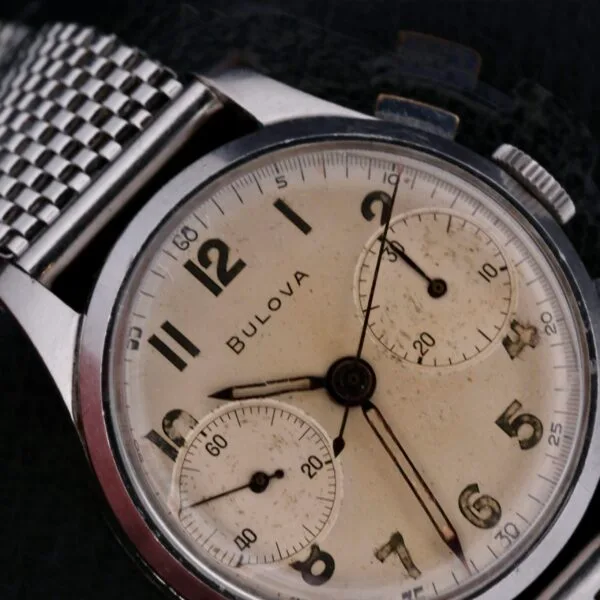 Close-up of a Bulova Chronograph 13AK Valjoux 23 Stainless Steel watch with a metal mesh band; it features a worn cream dial, black hour numbers, two subdials, and vintage-styled hands, emphasizing its classic design.