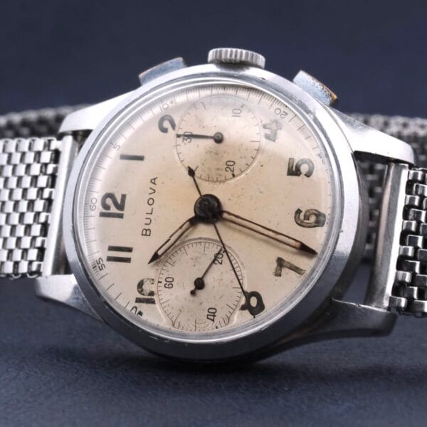 The Bulova Chronograph 13AK Valjoux 23 Stainless Steel wristwatch features a mesh strap, cream dial, three subdials, black Arabic numerals, and silver hands. Its case shows signs of wear that enhance its vintage appeal.