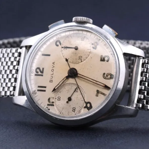 The Bulova Chronograph 13AK Valjoux 23 Stainless Steel wristwatch features a mesh strap, cream dial, three subdials, black Arabic numerals, and silver hands. Its case shows signs of wear that enhance its vintage appeal.