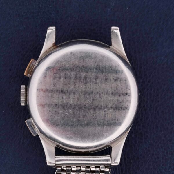 The image displays the back of a Bulova Chronograph 13AK Valjoux 23 Stainless Steel wristwatch with a metallic strap, set against a dark blue textured surface. The watch, featuring two side buttons, shows wear with scratches visible on its back.