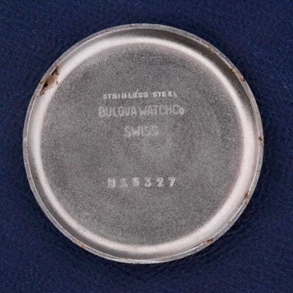 The silver circular watch case back is engraved with: STAINLESS STEEL BULOVA WATCH CO SWISS 915327. This Bulova Chronograph 13AK Valjoux 23 Stainless Steel features minor scuffs and tarnish, complementing a dark blue textured background.