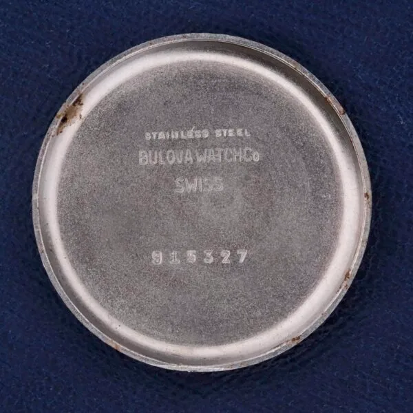 The silver circular watch case back is engraved with: STAINLESS STEEL BULOVA WATCH CO SWISS 915327. This Bulova Chronograph 13AK Valjoux 23 Stainless Steel features minor scuffs and tarnish, complementing a dark blue textured background.