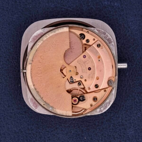 A close-up of the Omega Constellation TV Case Certified Chronometer movement reveals intricate gears, levers, and an engraved rotor on a dark textured surface, with metal components showcasing captivating gold and copper tones.