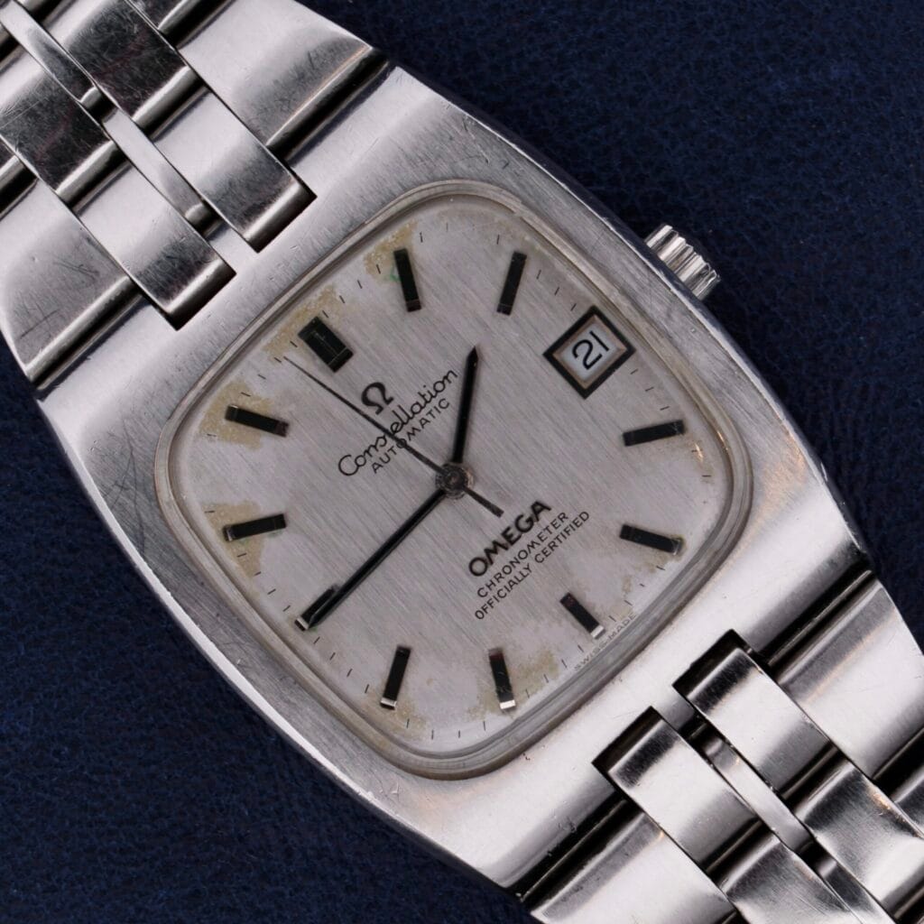 The Omega Constellation TV Case Certified Chronometer is a vintage automatic wristwatch with a silver square dial, date display, and a sleek metal band.